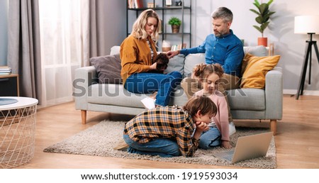 Nice Caucasian family with kids spending time at home. Little children boy and girl sitting on carpet floor watching videos or cartoons on laptop while mom and dad playing with pet puppy on couch