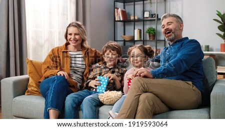 Caucasian joyful happy Caucasian family resting at home watching TV together sitting on couch with popcorn and laughing. Mom and dad with small son and daughter eating popcorn watching comedy movie