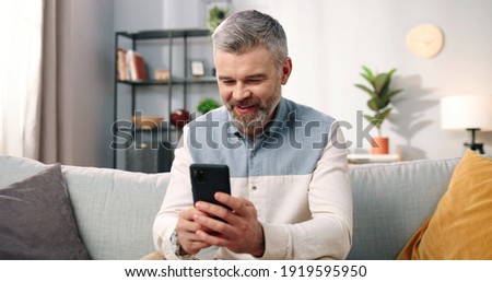 Close up of cheerful handsome middle-aged Caucasian man in positive mood texting on smartphone while sitting at modern home on sofa and smiling, grey-haired male using social network app on cellphone