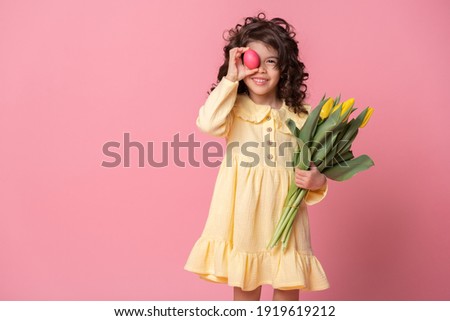 Funny child girl holding tulips and colorful Easter egg in front of her eye on pink background