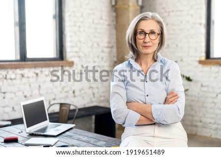 Serious and strict old senior business woman wearing smart casual shirt and stylish eyeglasses stands with arms crossed in modern office space. A mature bossy lady, elderly purposeful leader Royalty-Free Stock Photo #1919651489