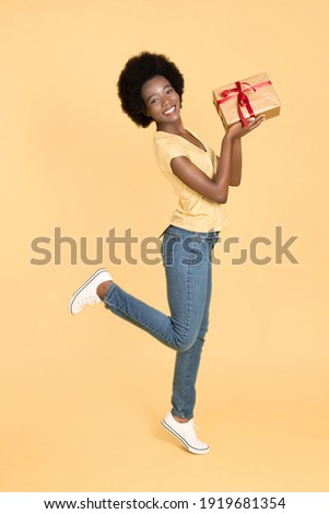 Full length body photo of excited funny black skinned woman with afro hair, wearing yellow t-shirt and denim jeans, posing umping with giftbox on isolated yellow background