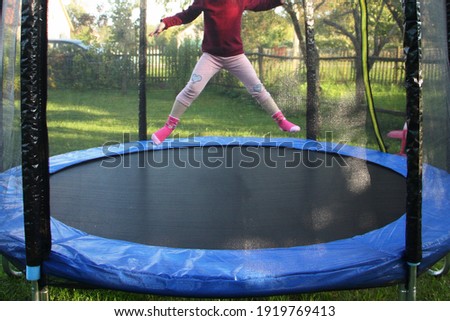 One little European girl jumps on a outdoor 8ft trampoline with outside safety soft net fence on green grass in the suburban Garden at Sunny summer day, active sport recreation Royalty-Free Stock Photo #1919769413