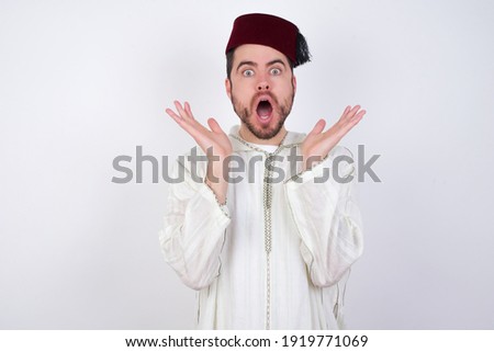 Surprised terrified young handsome Caucasian man wearing Arab djellaba and Fez hat over white wall Gestures with uncertainty, stares at camera, puzzled as doesn't know answer on tricky question.