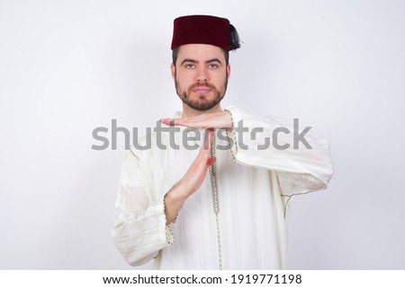 young handsome Caucasian man wearing Arab djellaba and Fez hat over white wall being upset showing a timeout gesture, needs stop, asks time for rest after hard work, demonstrates break hand sign