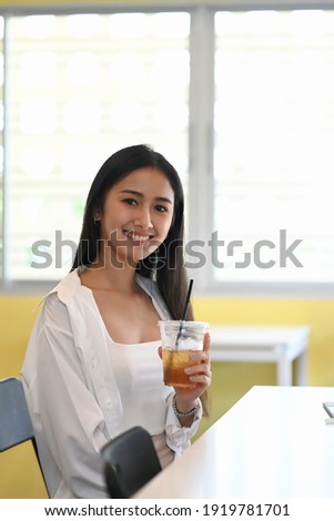 Portrait of cheerful young woman holding ice coffee and smiling to camera while sitting in office.