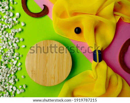 Abstract closeup top view round wooden mockup,pink green paper,white small pebble scattered on side,bright yellow semi-transparent folded wavy fabric,blue style napkin ring.Texture flatlay copy space.