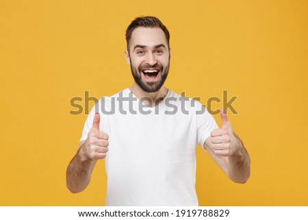 Excited funny young bearded man guy in white casual t-shirt posing isolated on yellow wall background studio portrait. People sincere emotions lifestyle concept. Mock up copy space. Showing thumbs up