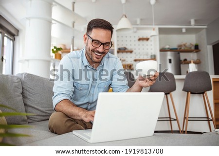 Young attractive man sitting on sofa at home working on laptop online, using internet, smiling, happy mood, freelancer, free leisure time, relaxed, modern job lifestyle