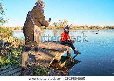 A mother takes pictures of her son in a hat and a red shirt is playing with the lake water on the pier