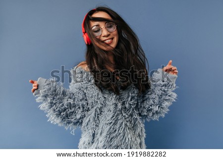 Wonderful lady with red headphones and round sunglasses in blue fashionable outfit playing her black long hair and smiling..