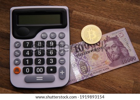 Hungarian forint 10,000 forint banknote Saint István King. Calculator on brown wooden table. Next to it is a gold bitcoin digital cryptocurrency coin and paper money. Bank image and photo.
