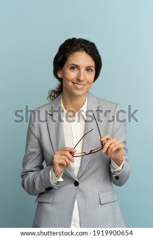 Confident businesswoman holding glasses, smiling, looking at camera posing isolated on blue background. Studio portrait of successful friendly female lawyer in blazer and blouse. Copy space.