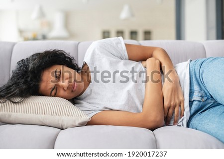 Sad young African American woman wearing casual clothes suffering from menstrual pain, feeling sick to her stomach, holding belly, having abdominal cramps during period and lying down on bed at home Royalty-Free Stock Photo #1920017237