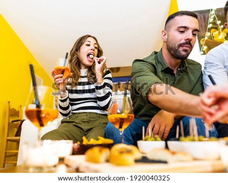 Young friends having fun at home aperitif with spritz cocktail and snacks on laden table. Selective focus on girl sitting on sofa eating mozzarella skewer holding a glass with orange long drink