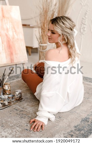 Beautiful girl artist sitting on the floor near the easel and paints. Blonde woman sitting in the artist's studio. 
Cute girl posing for artist.  Young female artist painting picture in studio.