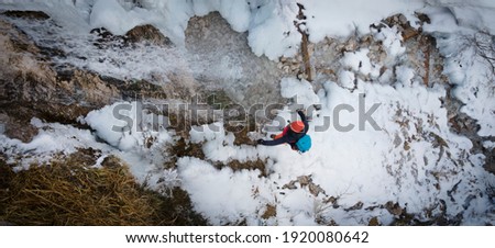 aerial drone shot of hiker with backpack, orange hat and orange pants standing next to waterfall surrounded by icicles and snow in winter