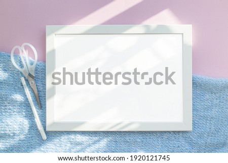 A blank layout of a white frame on an abstract pink table and a blue knit fabric. The light from the sun makes patterns. The concept of minimalism and modernity. White pen and scissors.