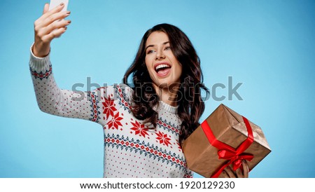Cheerful woman with gift box taking selfie with smartphone on blue background