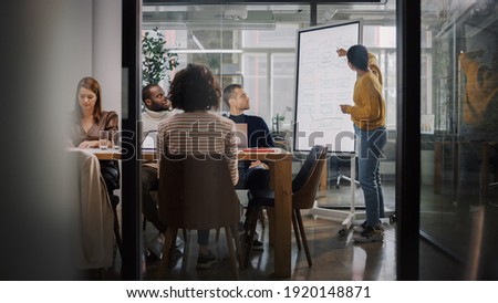 Project Manager Makes a Presentation for a Young Diverse Creative Team in Meeting Room in an Agency. Colleagues Sit Behind Conference Table and Discuss Business Development, User Interface and Design. Royalty-Free Stock Photo #1920148871