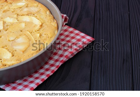 freshly made apple pie in a baking dish. Stands on a red-white napkin in a cage on a black, wooden background. Homemade baking concept. High quality photo