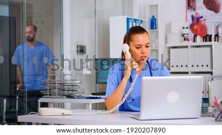 Medical staff talking with patient on phone from hospital about diagnosis, male nurse working in background. Healthcare physician, receptionist doctor assistant helping with telehealth communication