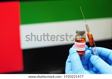 COVID-19 vaccine. Hands in blue medical gloves holding a vaccine bottle and syringe with United Arab Emirates flag as background