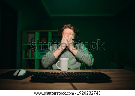 Gamer in a headset sleeps at night in a room with a green light and covers his face with his hands. Frustrated gamer hides from losing at hand.