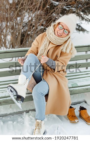 young woman in Park near the ice skating rink. girl in beige coat and hat is going to skate, winter leisure and relaxation, tying shoelaces on ice skates before skating on the ice rink

