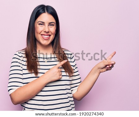 Young beautiful brunette woman wearing casual striped t-shirt over isolated pink background smiling and looking at the camera pointing with two hands and fingers to the side.