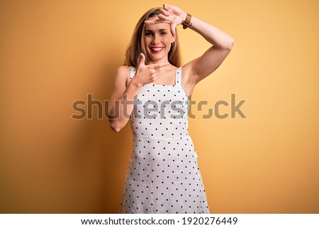 Young beautiful blonde woman on vacation wearing summer dress over yellow background smiling making frame with hands and fingers with happy face. Creativity and photography concept.
