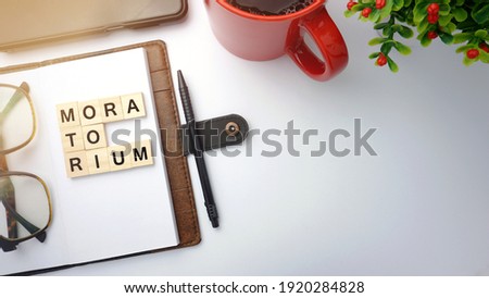 Selective focus on smartphone, pen, diary, red cup, artificial flower and wood blocks with word MORATORIUM on top of table