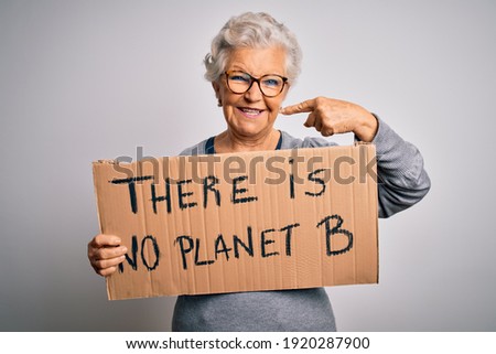 Senior beautiful grey-haired woman asking for environment holding banner with earth message with surprise face pointing finger to himself