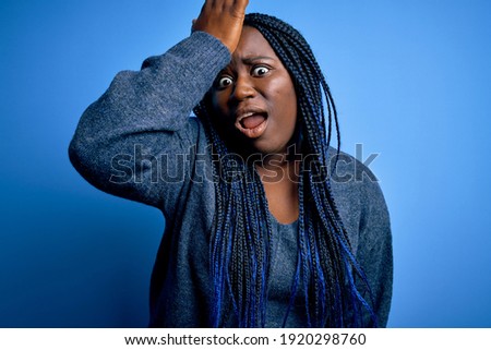 African american plus size woman with braids wearing casual sweater over blue background surprised with hand on head for mistake, remember error. Forgot, bad memory concept.