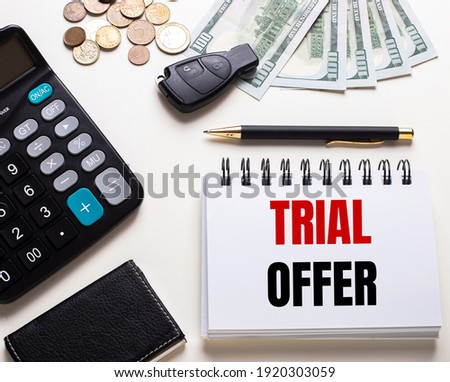 On a white table is a calculator, car key, cash, a pen and a notebook with the inscription TRIAL OFFER
