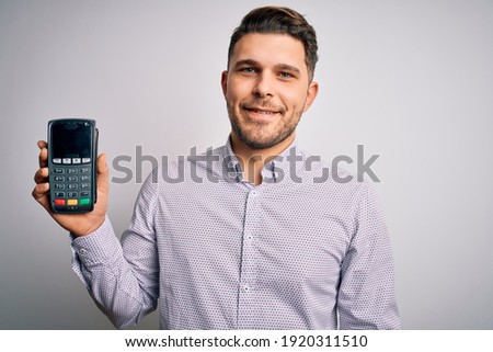Young business man with blue eyes holding dataphone payment terminal over isolated background with a happy face standing and smiling with a confident smile showing teeth