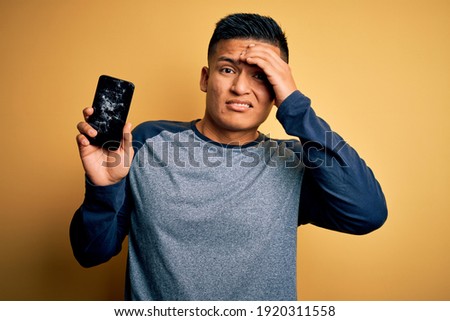 Young handsome latin man holding broken smartphone showing craked screen stressed with hand on head, shocked with shame and surprise face, angry and frustrated. Fear and upset for mistake.