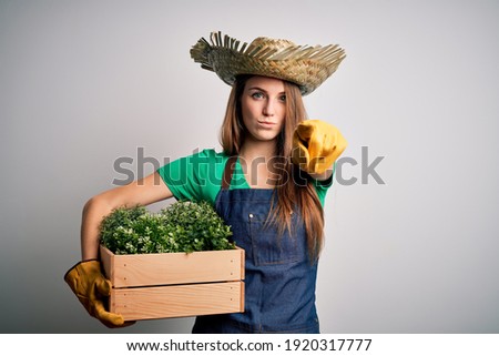 Young beautiful redhead farmer woman wearing apron and hat holding box with plants pointing with finger to the camera and to you, hand sign, positive and confident gesture from the front