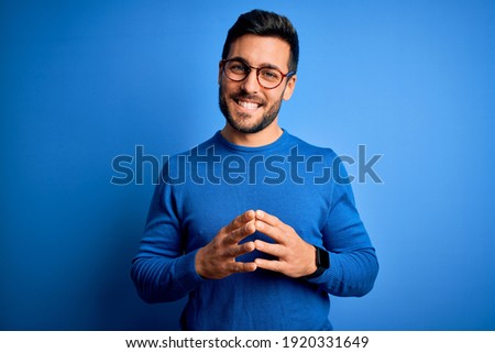 Young handsome man with beard wearing casual sweater and glasses over blue background Hands together and fingers crossed smiling relaxed and cheerful. Success and optimistic Royalty-Free Stock Photo #1920331649