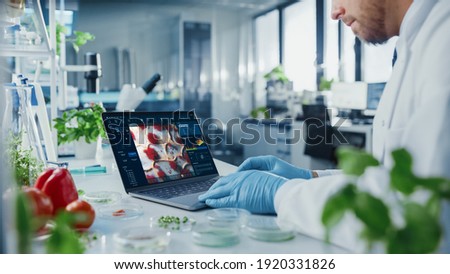 Male Scientist Working on a Laptop Computer with Display Showing Gene Editing Interface. Microbiologist is Rubber Gloves in a Bright Modern Food Laboratory with Advanced Technological Equipment.