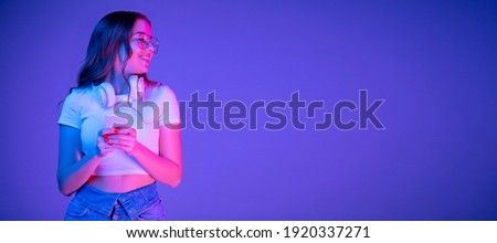 Caucasian woman's portrait on blue background in multicolored neon light. Beautiful model with headphones, phone. Concept of human emotions, facial expression, sales, ad, fashion. Copyspace. Flyer