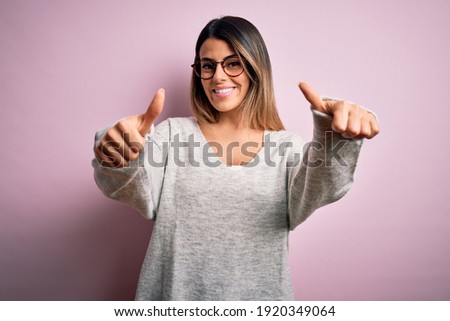 Young beautiful brunette woman wearing casual sweater and glasses over pink background approving doing positive gesture with hand, thumbs up smiling and happy for success. Winner gesture.