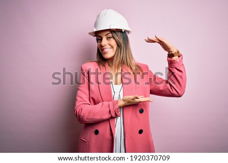 Young beautiful brunette architect woman wearing safety helmet over pink background gesturing with hands showing big and large size sign, measure symbol. Smiling looking at the camera. Measuring