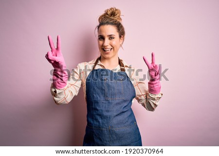 Young beautiful blonde cleaner woman doing housework wearing arpon and gloves smiling looking to the camera showing fingers doing victory sign. Number two. Royalty-Free Stock Photo #1920370964
