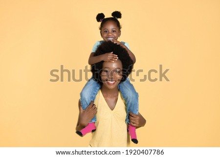 Joyful smiling young black woman mother giving her adorable pretty daughter piggy back ride, isolated on yellow with copy space