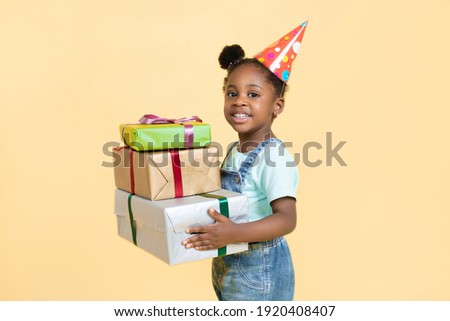 happy child smiling african girl, wearing birthday hat and casual jeans overalls, holding stack og colorful gift boxes, stands on isolated yellow background