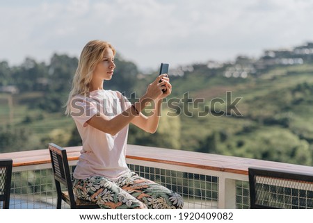 Blonde woman sitting in outdoor cafe and take picture of amazing view
