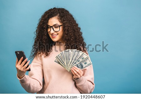 Happy winner! Portrait of a cheerful young curly woman holding money banknotes and celebrating isolated over blue background. Using phone.