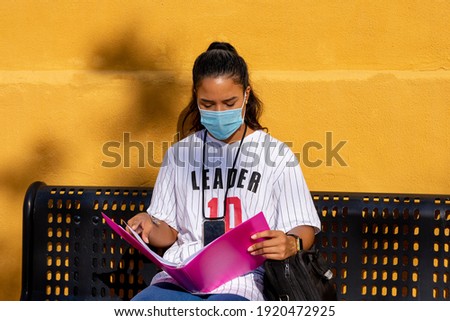 Afro-Ecuadorian college girl with a white t-shirt and jeans sitting on a bench studying with a pink folder on a yellow background