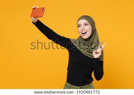 Funny young arabian muslim woman in hijab black green clothes doing selfie shot on mobile phone showing victory sign isolated on yellow background studio portrait. People religious lifestyle concept
